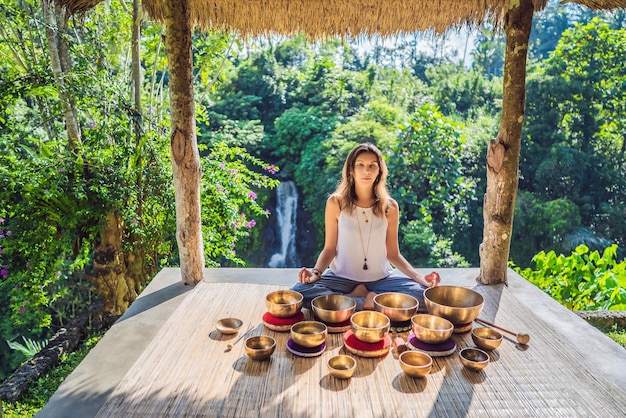 Photo woman playing on tibetan singing bowl while sitting on yoga mat against a waterfall vintage tonned beautiful girl with mala beads meditating