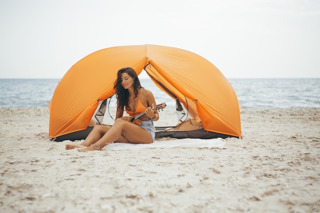 Woman playing the ukelele with an orange tent on the beach
