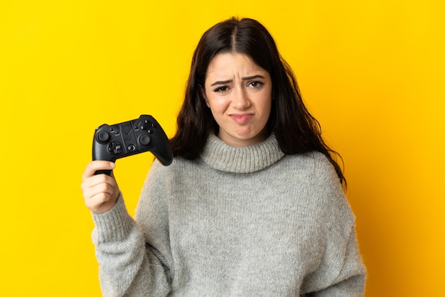 Женщина играет с контроллером видеоигры, стоя на желтой стене с грустным выражением лица
