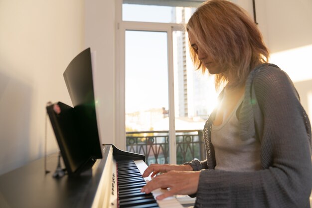 Una donna suona il pianoforte elettronico sullo sfondo di una grande finestra attraverso la quale penetra la luce del sole