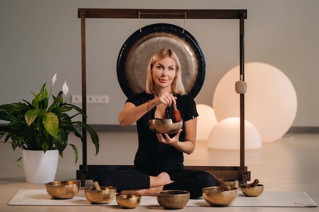 A woman plays a Tibetan singing bowl while sitting on a yoga mat against the background of a gong