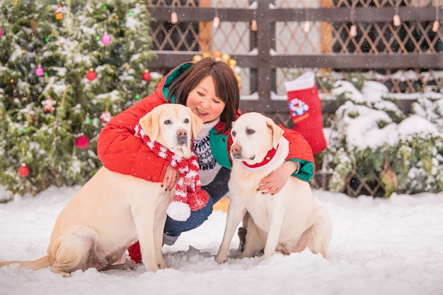 A woman plays with labradorsmi dogs near a decorated Christmas tree during a snowfall in winter in the courtyard of an apartment building.