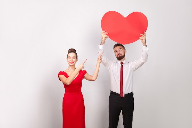 Woman pointing finger at big heart, man looking at heart. Indoor, studio shot, isolated on gray background