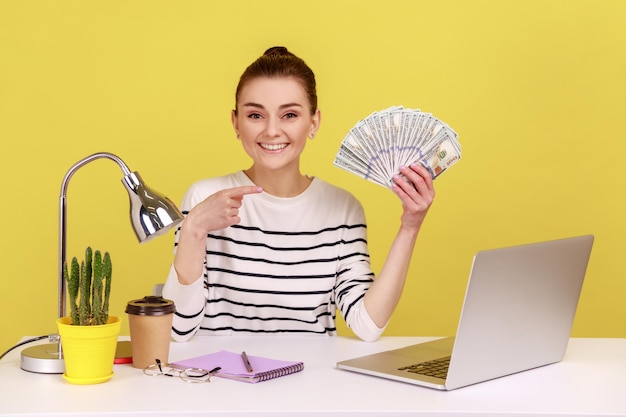 Photo woman pointing finger at hundred dollar bills holding sitting at workplace with laptop high salary