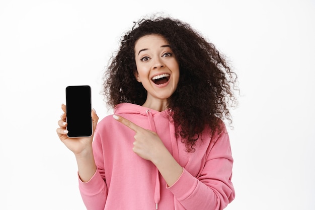 woman pointing finger at smartphone empty screen standing on white