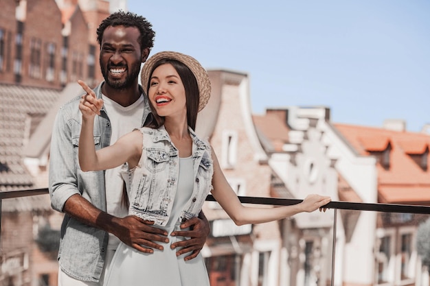 Woman pointing with hand on something while man hugging from beside standing on glass balcony with old buildings on the background