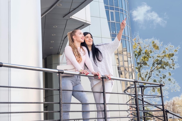 woman points a finger up to her friend while standing behind a railing at a large glass building in the city.