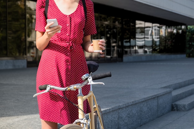 Woman in polka dot dress with  bike commuting, checking mail online and having coffee. Female person using smartphone and holding cup of hot drink on her way to work