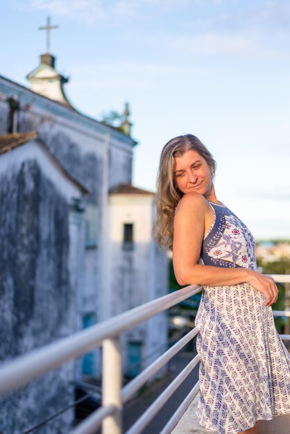 A woman on the porch of her house in light clothes looking at\
the street against the sky and church in the background