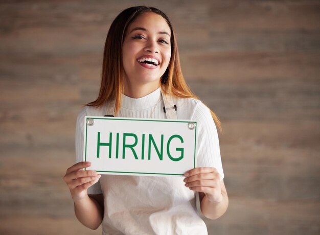 Woman portrait and hiring sign for small business recruitment career or job opportunity against a studio background Happy female entrepreneur with apron holding billboard poster for hire on mockup