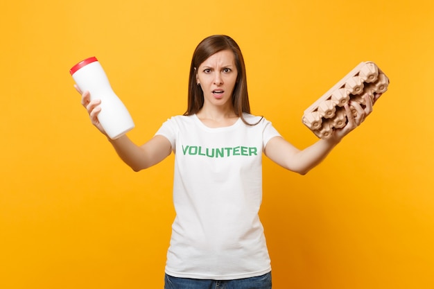 Woman portrait in white t-shirt with written inscription green title volunteer hold plastic bottle, cardboard box isolated on yellow background. Voluntary free assistance help, trash sorting concept.