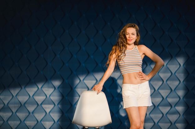 Woman poses before a white chair on blue background