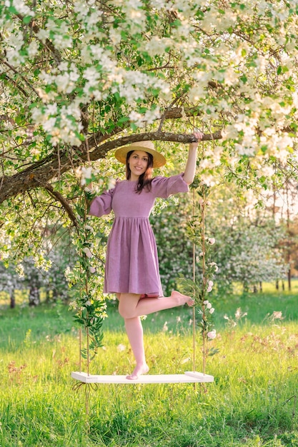 A woman poses standing on a rope swing in a blooming apple\
orchard