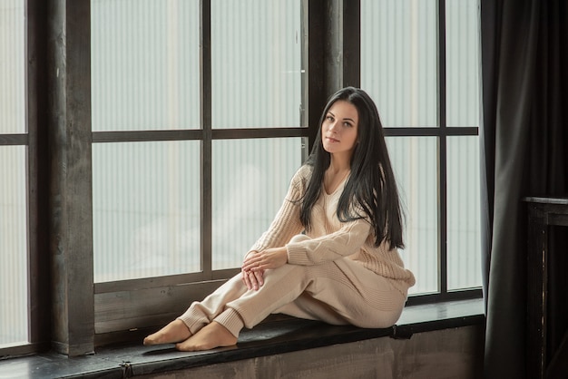 Woman posing in fashionable clothes near the window. Woman at the window in a beige knitted suit.