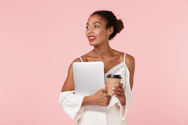 Woman posing isolated over pink wall looking aside drinking coffee holding laptop computer