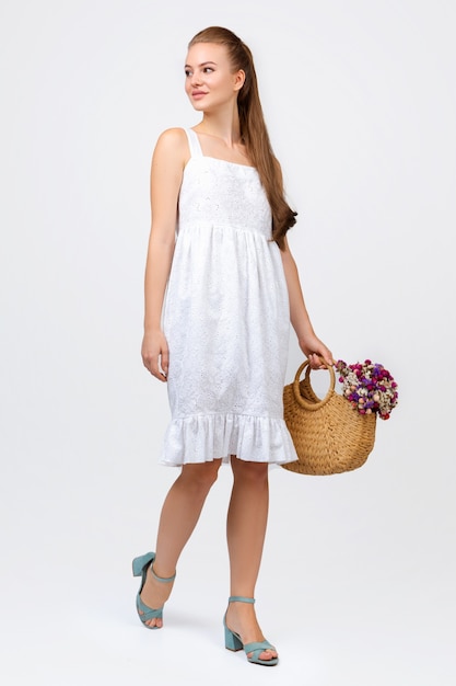 Woman posing on white wall in a white dress with a handbag and flowers