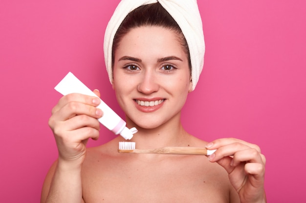 Woman posing with bare sgoulders and white towel on head, putting some toothpaste on toothbrush