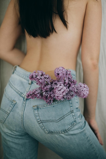 Woman posing with lilac flowers in denim jeans pocket on rustic
background blooming lilac flowers in back pocket creative image
sensual atmospheric mood hello spring