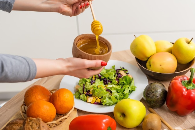 Woman pouring honey mustard dressing into bowl with fresh salad on table.