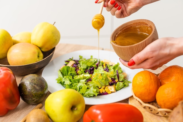 Woman pouring honey mustard dressing into bowl with fresh salad\
on table.