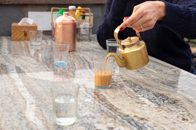 Woman pouring masala tea from teapot into cupglass on table in cafe.