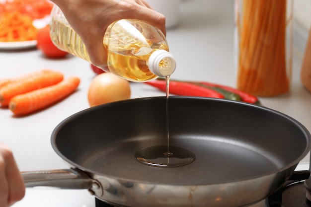 Женщина заливает масло из бутылки в кастрюлю на кухне, рядом свежие овощи и макароны.