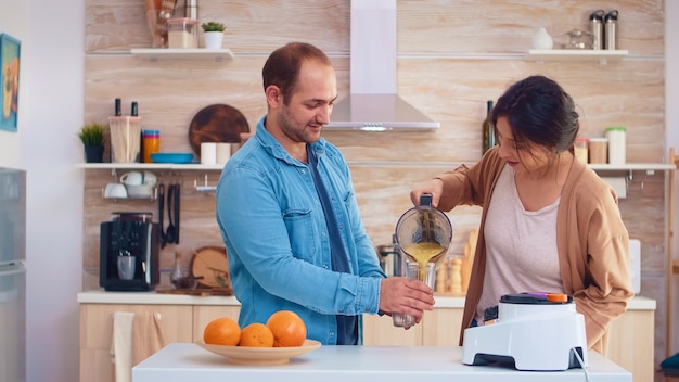 Woman pouring tasty smoothie from blender in glasses for her and husband. Cheerful family making together organic healthy fresh nutritious tasty juice for breakfast from fresh fruits while on a diet.