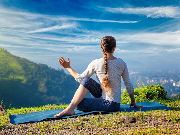 Woman practices yoga asana Parivrtta Marichyasana seated spinal twist outdoors in mountains