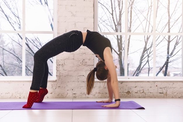 Йога женщины практикуя делая представление Ardha Chakrasana в светлой комнате на циновке йоги внутри помещения. Разогреть,