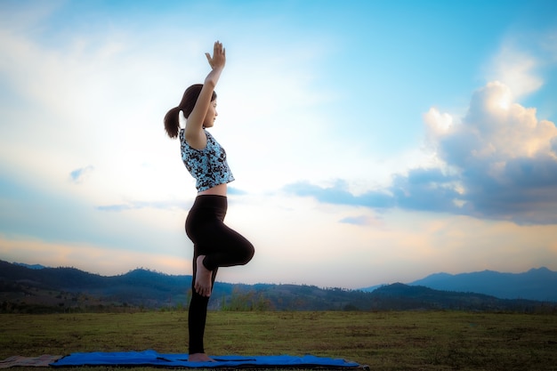 Woman practicing yoga pose outdoors over sunset sky background.