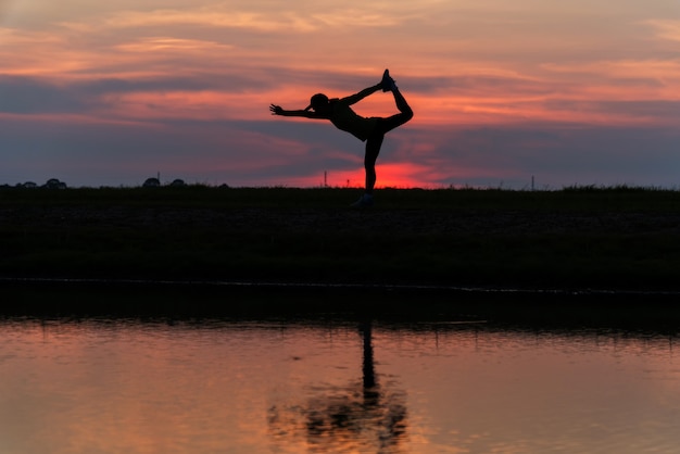 Woman practicing yoga during surrealistic sunset at the seaside. healthy concept and workout. Silhouette asian woman exercise yoga pose at twilight sunset time with reflection on river.