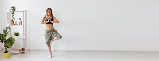 Woman practise yoga performs tree pose standing inside of cozy room with plants and greenery healthy