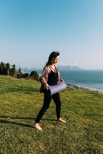 Woman prepares to place her yoga mat on the grass in front of the sea. Copy space