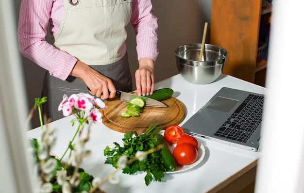 女性は、レシピを見ながらラップトップ コンピューターを使用してキッチンで野菜サラダを準備します。