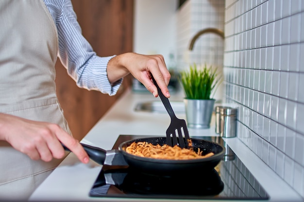 モダンなロフトスタイルのキッチンで夕食のためにストーブのフライパンで食事を準備する女性