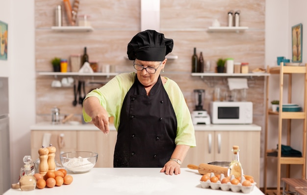 Photo woman preparing tasty pizza sprinkling baking powder on kitchen table. happy elderly chef with uniform sprinkling, sieving sifting raw ingredients by hand.