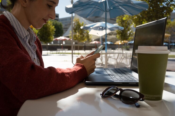Woman in profile smiling working outdoors with laptop and looking at cell phone, enterprising woman concept, with copy space.