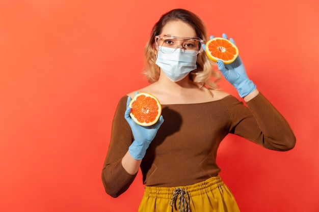 Woman in protective glasses mask gloves holding orange fresh fruit natural vitamins for immune system and flu treatment healthy nutrition during quarantine indoor studio shot red background