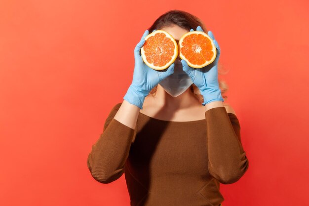 Foto donna in guanti protettivi e maschera facciale che copre gli occhi con frutta fresca arancione, vitamine naturali per il sistema immunitario e trattamento dell'influenza, alimentazione sana durante la quarantena. girato in studio, isolato