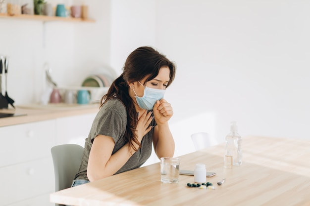 Woman in protective mask coughing in fist sick of coronavirus viral infection spreading coronavirus covering mouth and nose. Painful cough ill patient sitting in kitchen at home quarantine