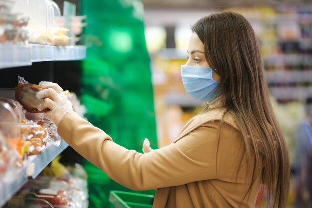 Woman in protective mask and gloves reading label with price at goods while standing in supermarket