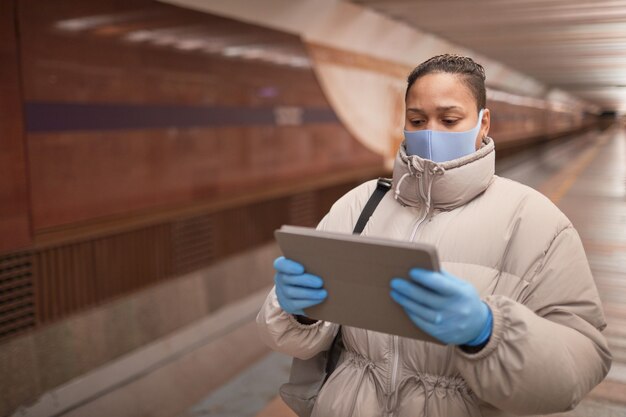 Woman in protective mask and gloves using digital tablet while walking along in the underground