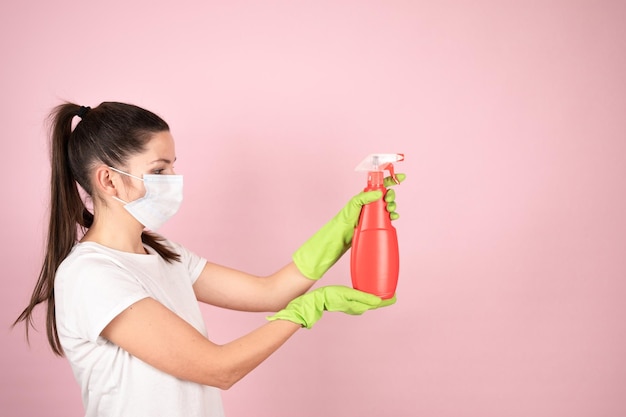 Donna in maschera protettiva e guanti verdi con flacone spray chimico su sfondo rosa concetto di prodotti per la pulizia