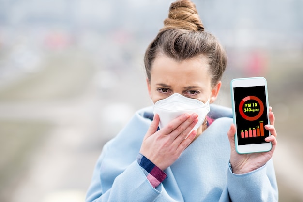 Donna in maschera protettiva che tiene in mano uno smartphone con misurazione dell'inquinamento atmosferico di pm10 all'aperto