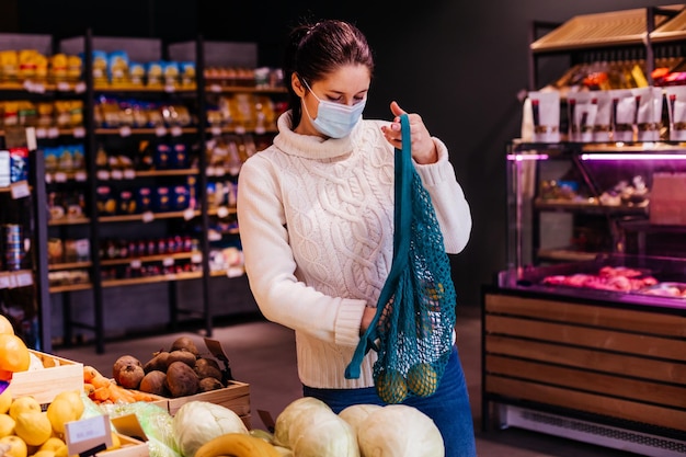 Woman in protective mask is choosing fruits and vegetables at food market putting them inside her blue shopping mesh bag Reusable eco bag for shopping Zero waste while pandemic concept