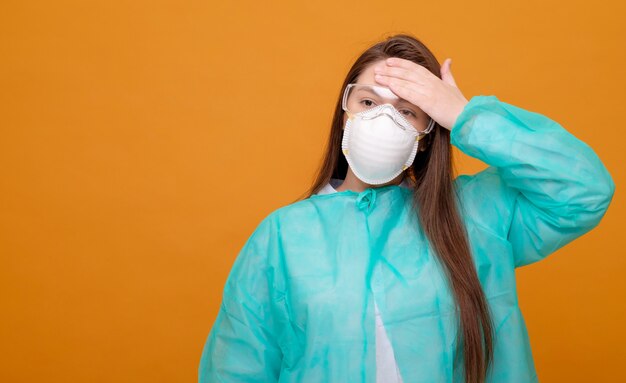 Woman in protective medical equipment with medical mask on face on yellow symptom of illness coronavirus pandemic headache