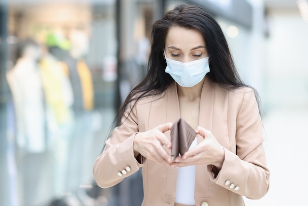 Woman in protective medical mask looks into empty wallet in mall