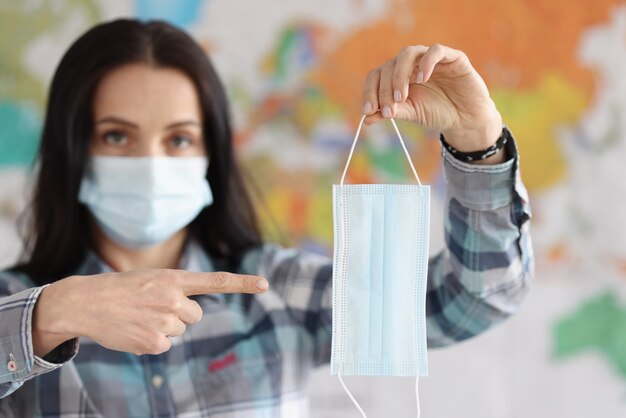 Woman in protective medical mask points at medical mask with her finger against background of world map