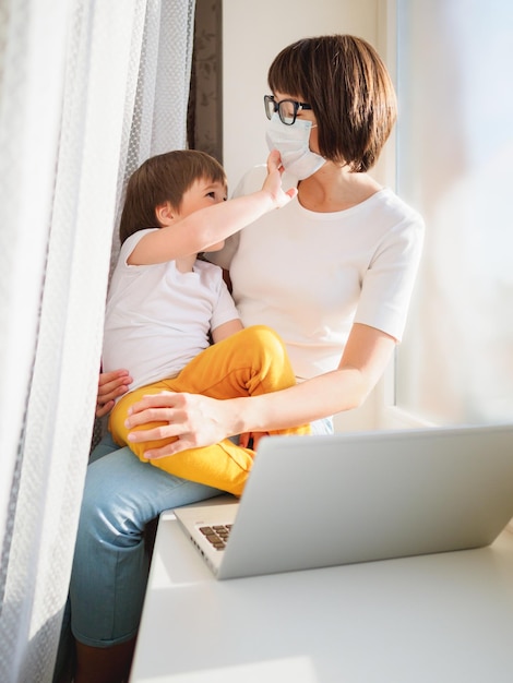 woman in protective medical mask works remotely from home with laptop and boy coronavirus covid19