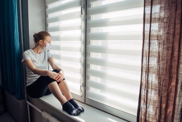 Woman in protective medical masks sits on windowsill and looks out window